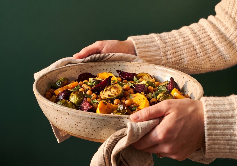Hands carrying a stoneware dish of roasted vegetables to a potluck